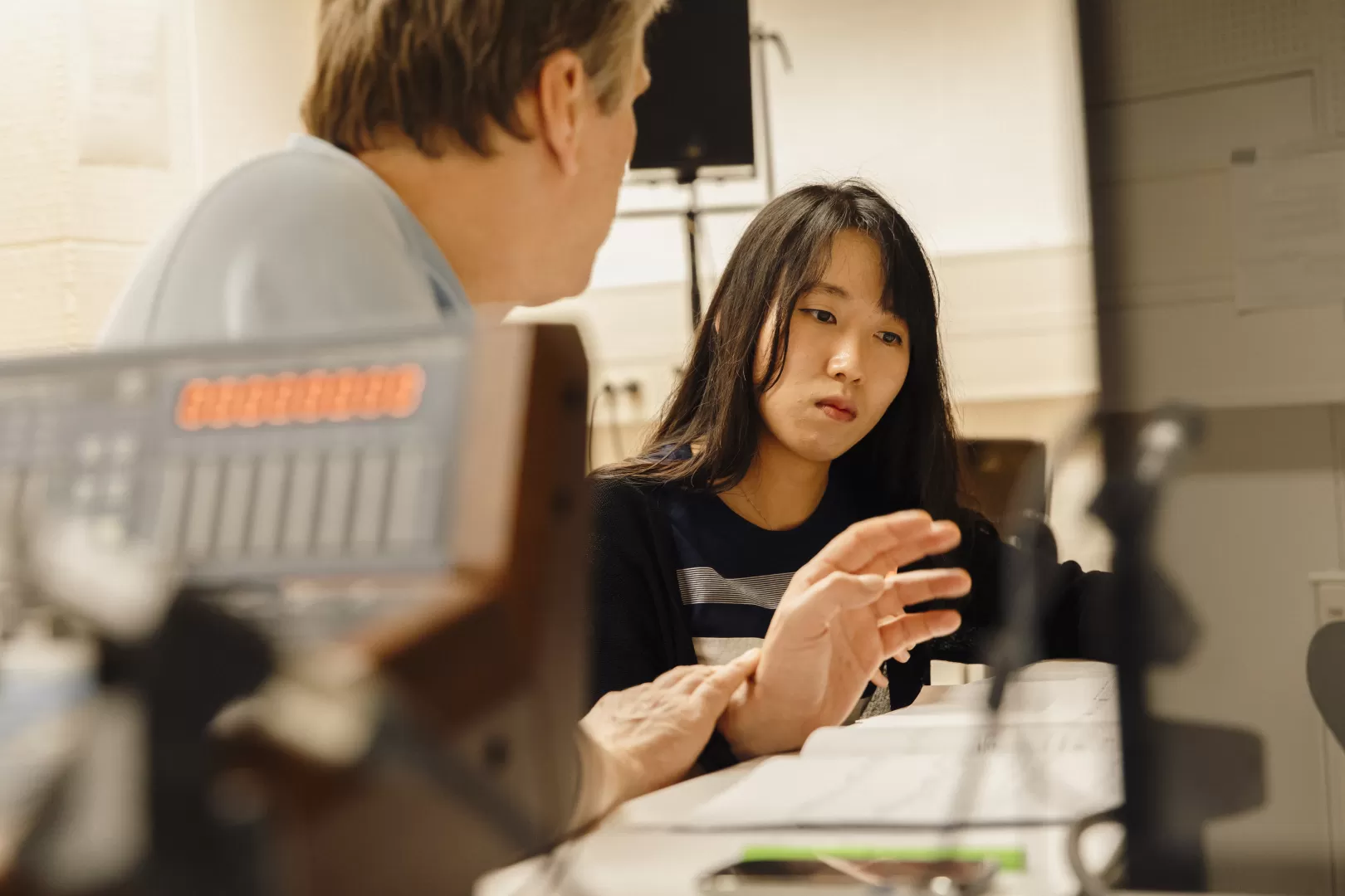 Eine junge Frau und ein Mann, der unterrichtet sitzen am Tisch, die Frau hört dem Unterricht zu.