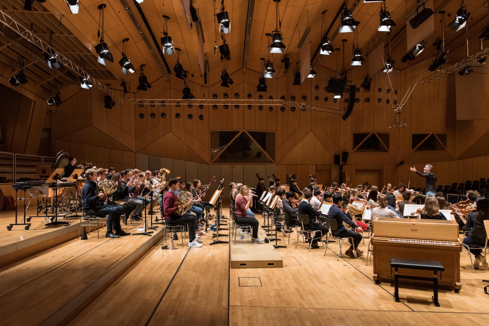 Seitliche Ansicht eines Orchesters auf der Bühne, rechts im Bild der Dirigent.