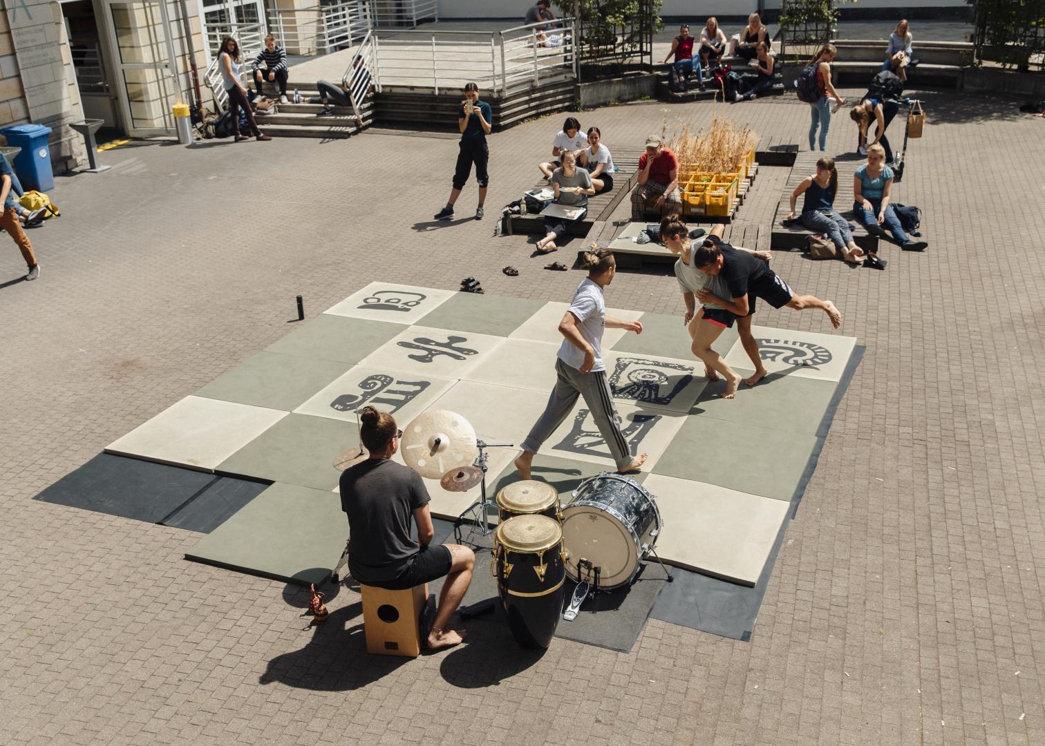 Sommer im Innenhof der HfMDK, Studierende tanzen, sitzen in der Sonne, eine Person spielt Schlagzeug.