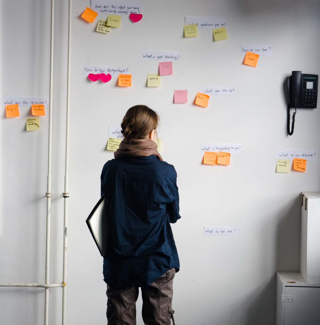 Eine Frau betrachtet eine Wand, an der Post-Its mit Fragen kleben.