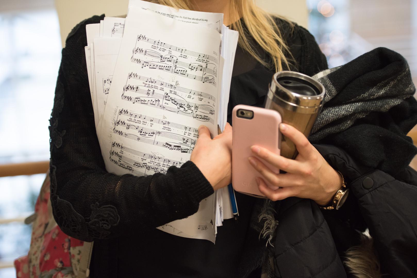 Eine Studentin hält Noten, einen Kaffeebecher, eine Jacke und ihr Smartphone in der Hand.