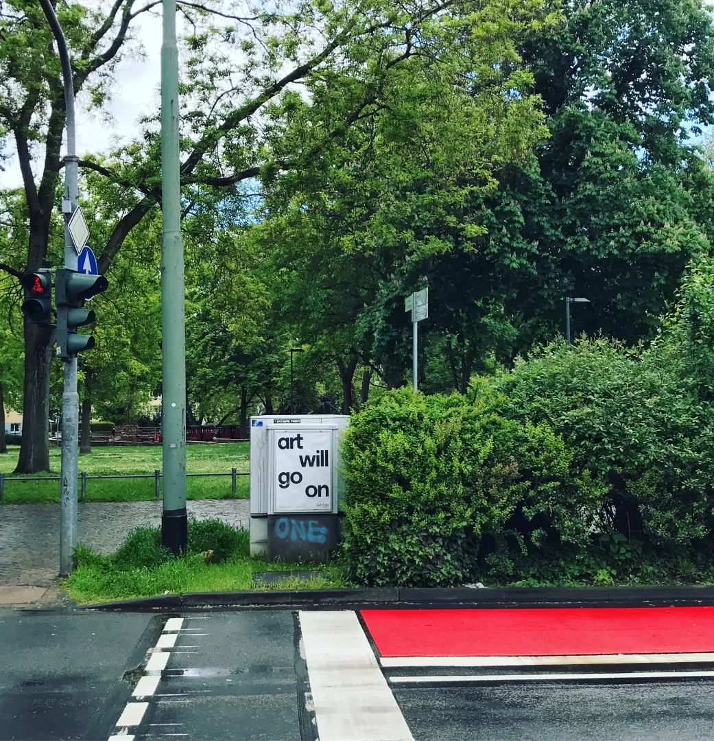 Straße in Frankfurt mit roten Radweg, an einem Stromkasten hängt ein Plakat der HfMDK mit der Aufschrift "Art will go on"