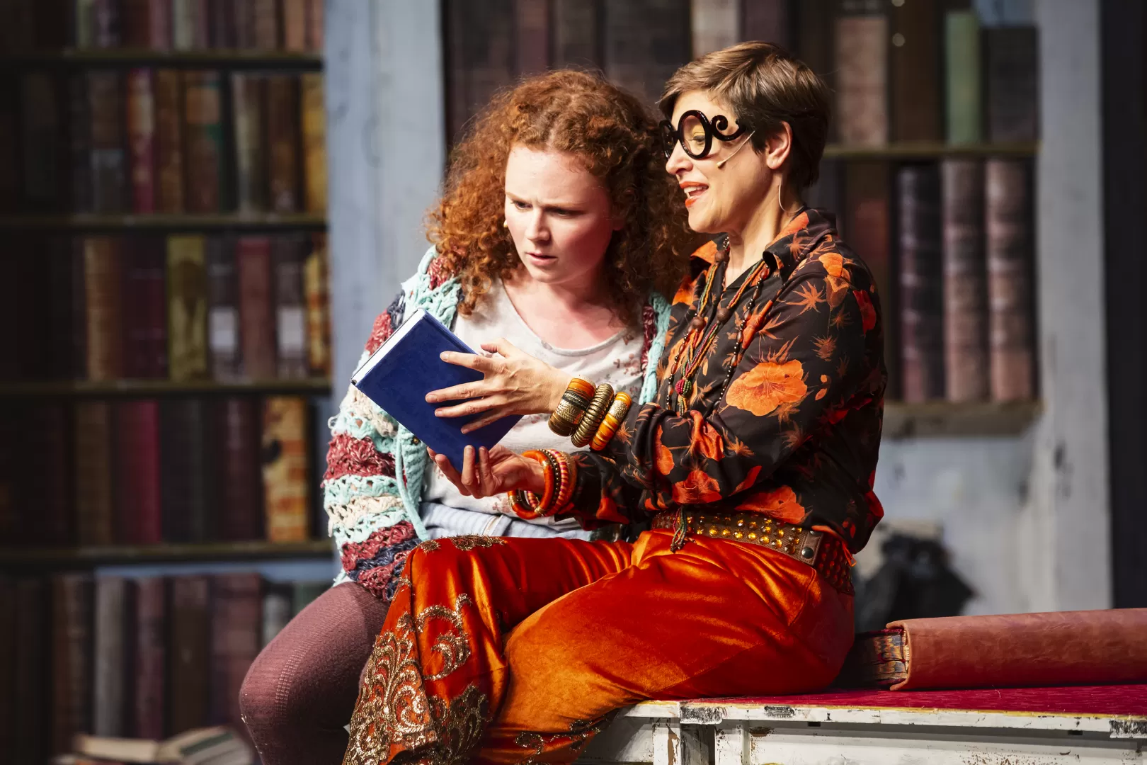 Zwei Schauspielerinnen sitzen auf einem Tisch vor großen Bücherregalen und schauen sich ein blaues Buch an.
