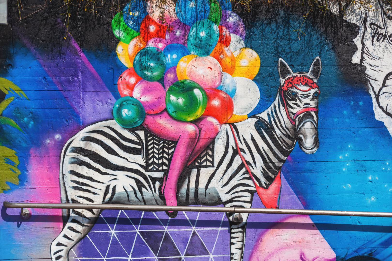 Buntes Graffiti im Stadtbild: Eine Person auf einem Zebra, die Arme übervoll mit bunten Luftballons.