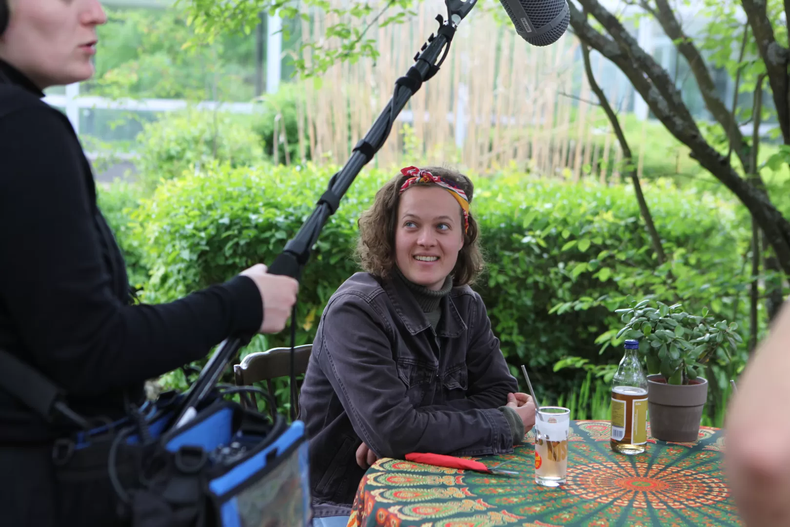 Schauspielstudentin sitzend an Tisch mit gemusterter Tischdecke und Blick zur Tonfrau am Bildrand.
