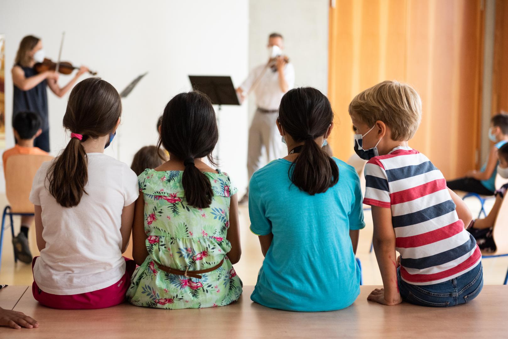 Vier Kinder sitzen mit dem Rücken zum Betrachter und hören zwei Musikern zu.