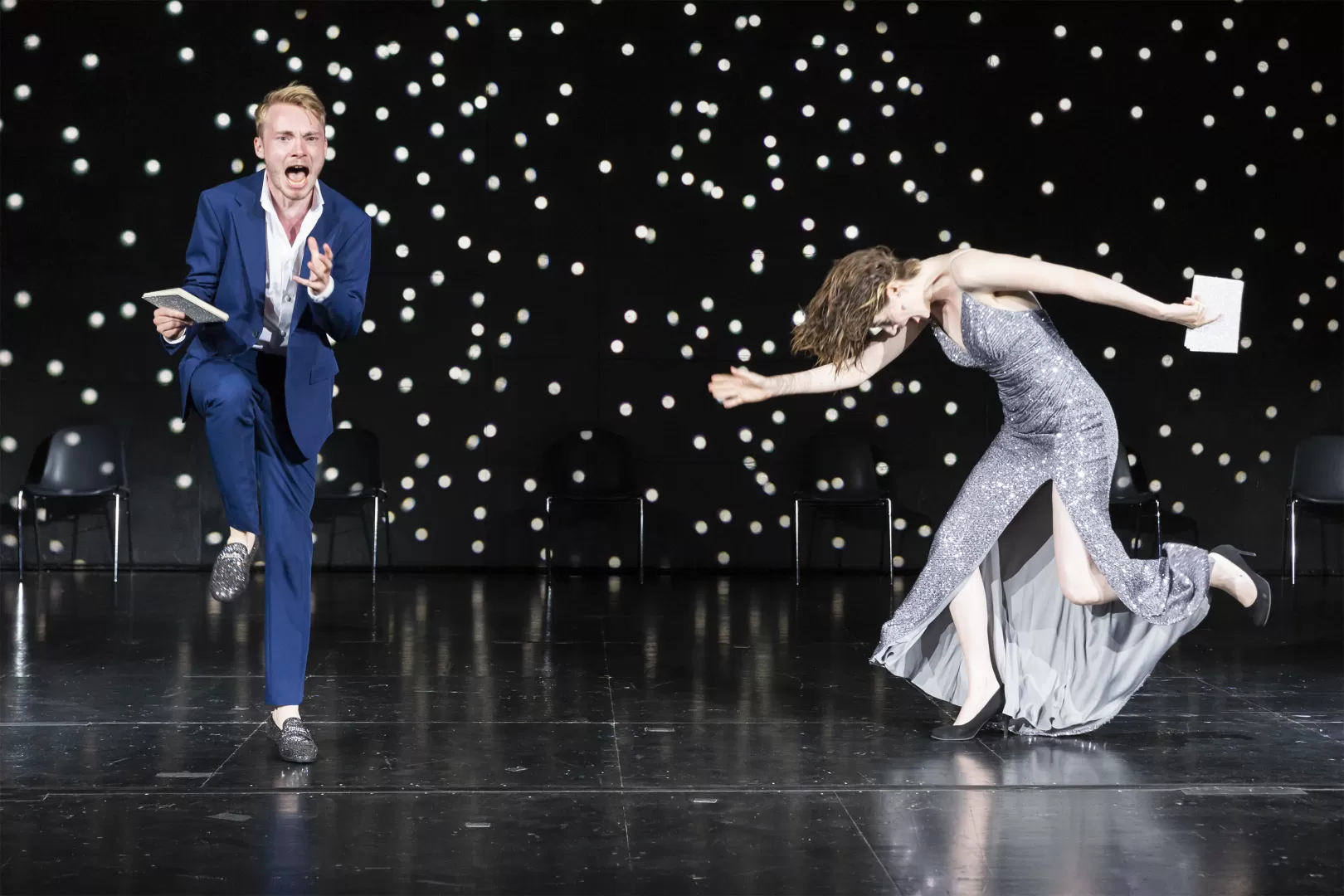 Schauspieler in blauem Anzug, dynamische Bewegung, und Schauspielerin in silbernem Glitzerkleid, seitlich und in Bewegung.