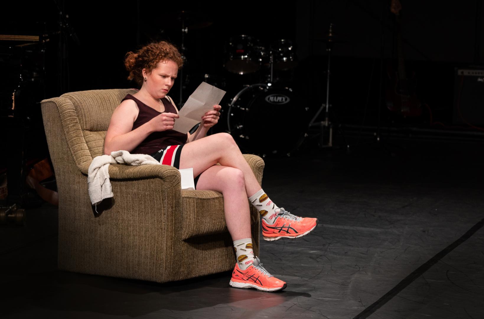 Schauspielerin in Sportbekleidung sitzt in einem altmodischen Sessel und liest kritisch ein Papier.
