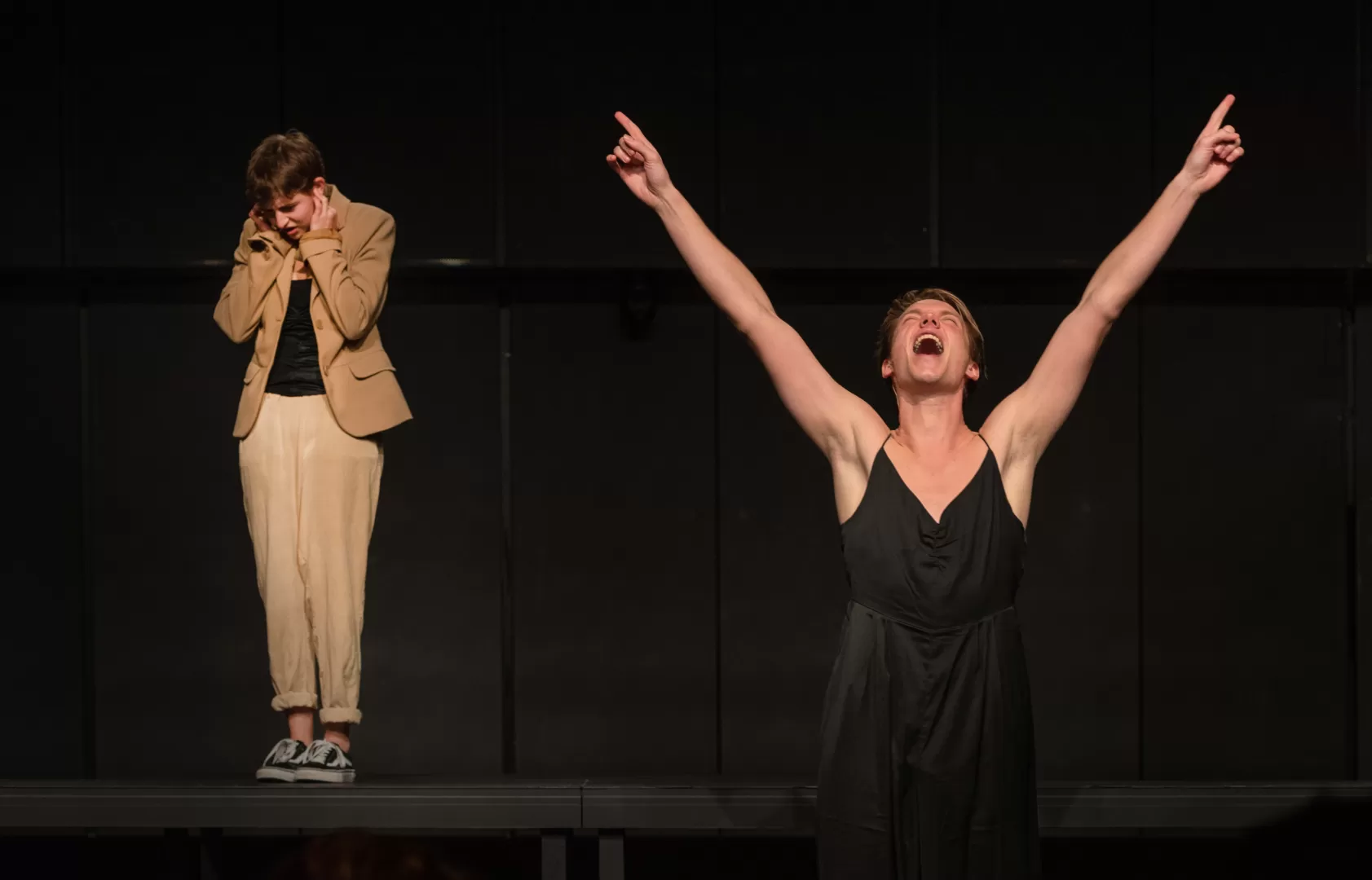 Schauspieler, vorne, mit nach oben ausgestreckten Armen. Im Hintergrund eine Schauspielerin, die sich die Ohren zuhält.