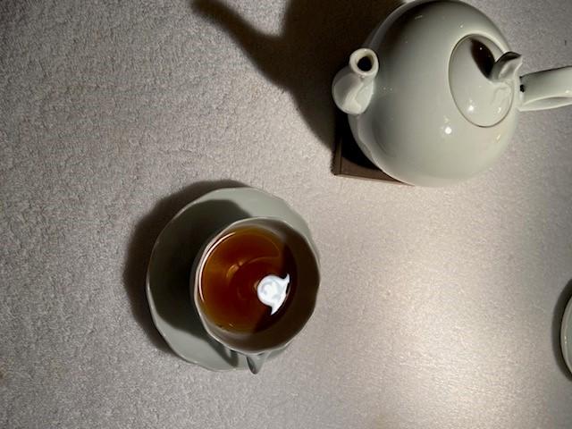 Aus der Vogelperspektive ist auf einem weissen Untergrund eine weiße Porzellankanne Tee oder Kaffee und links daneben eine weiße Tasse mit einer dunklen Flüssigkeit und einem weissen Tupfer Milch.