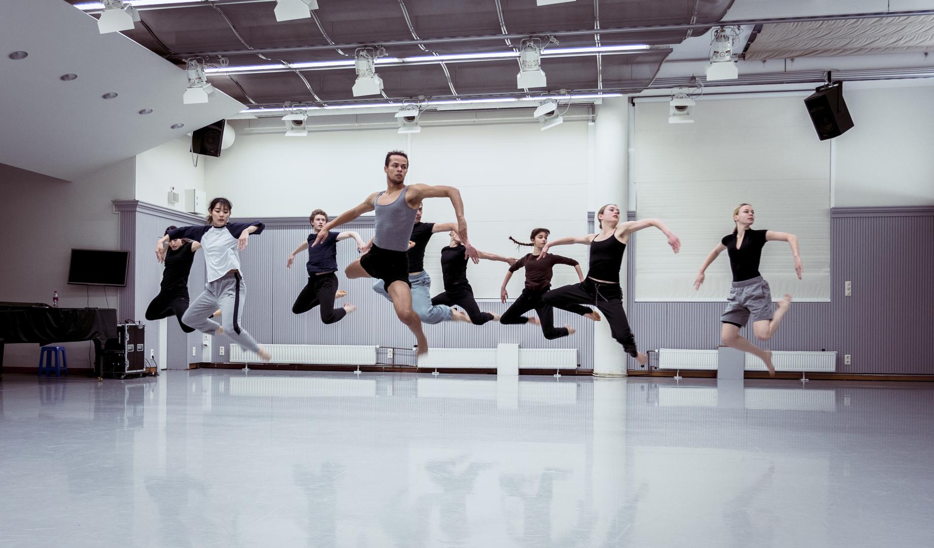 Neun Tänzerinnen und Tänzer beim Training im Tanzstudio, fotografiert mitten im Sprung.