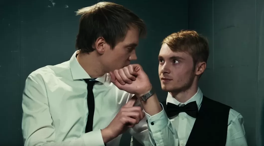 Zwei Männer im Fahrstuhl. Der eine zeigt auf seine Armbanduhr.