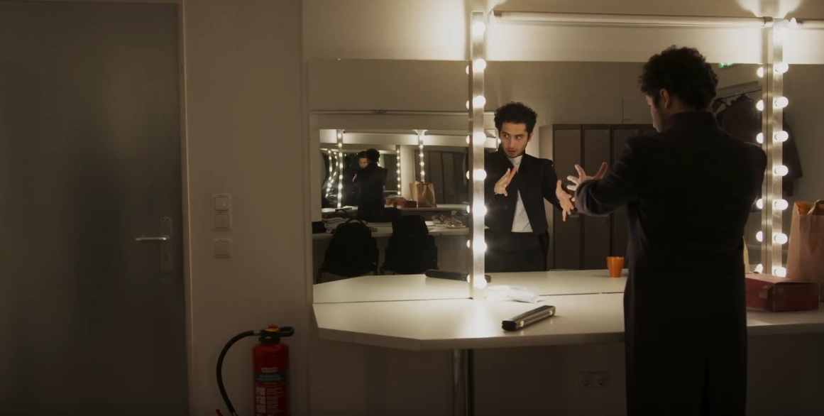 Ein Dirigent steht nervös und mit ausgestreckten Armen vor dem Spiegel in seiner Garderobe.