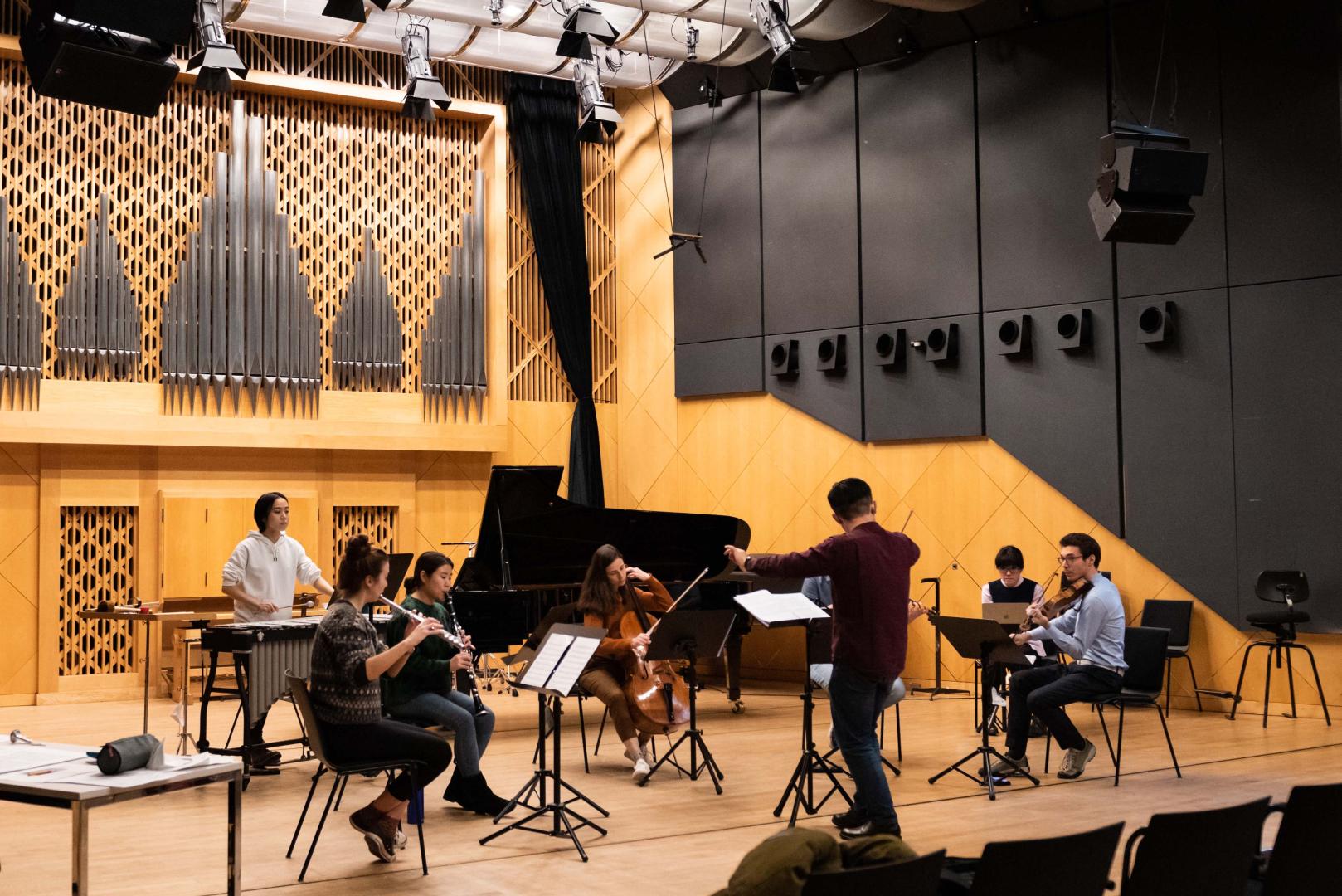 Ensemble mit 7 Musiker*innen auf der Bühne, im Vordergrund sieht man den Dirigenten von hinten.