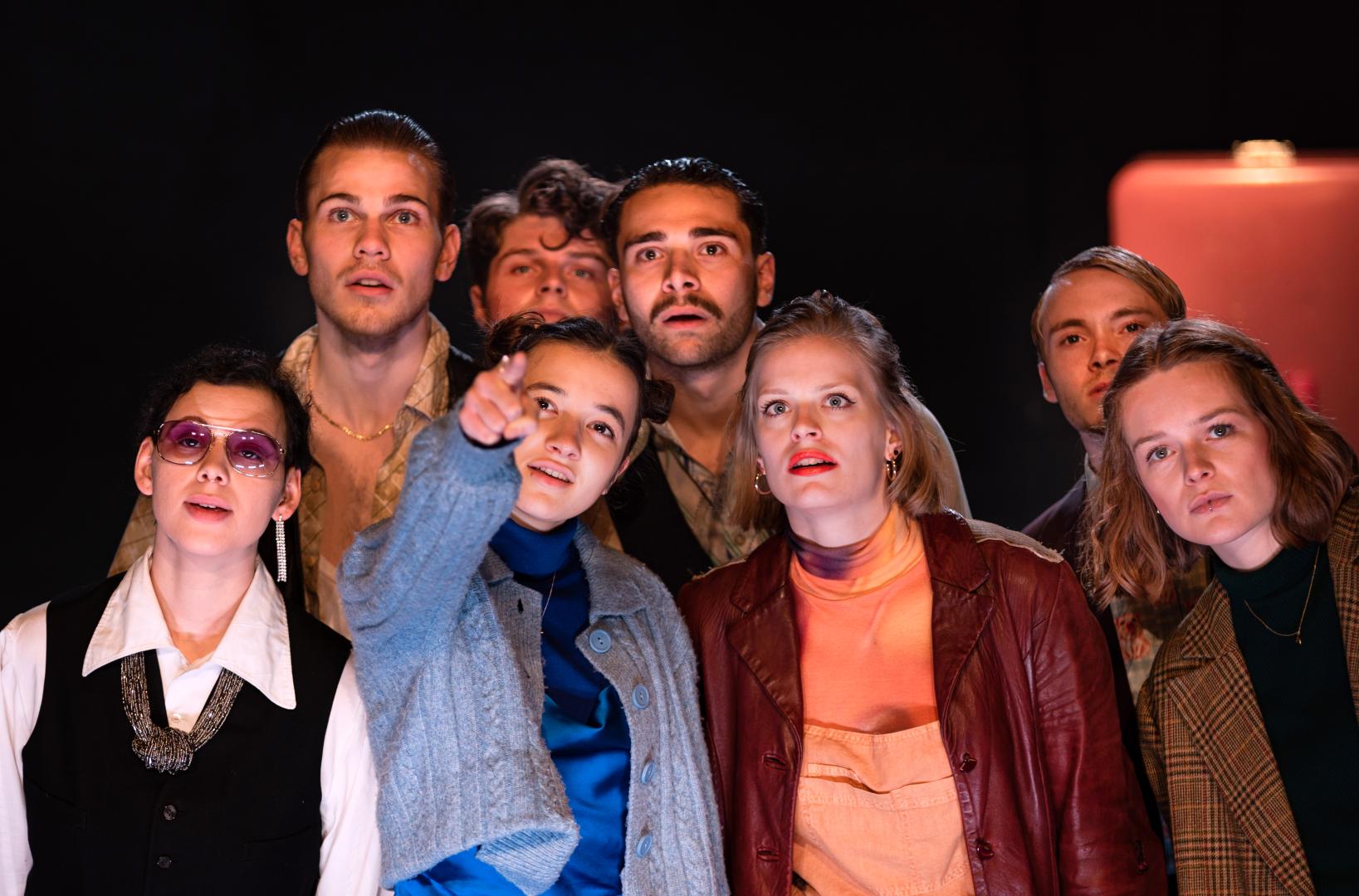 Gruppenfoto eines Schauspiel-Ensembles, eine Schauspielerin deutet in Richtung Kamera, alle folgen ihrem Blick.