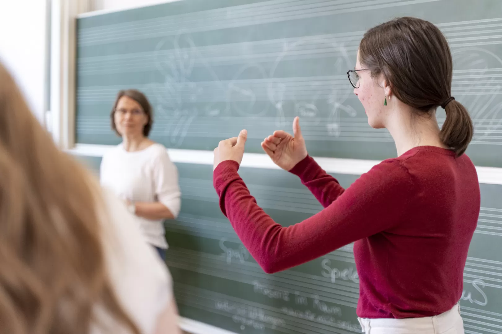 Eine Studentin im roten Pulli steht vor einer Tafel mit Notenlinien und erläutert etwas, dabei gestikuliert sie mit den Händen.