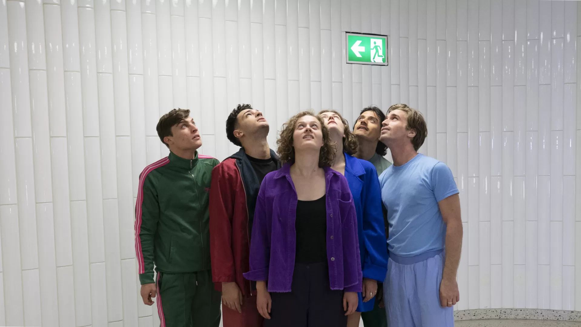 Sechs Schauspielabsolvierende nebeneinander, den Blick nach oben zu einem Notausgangsschild gerichtet.