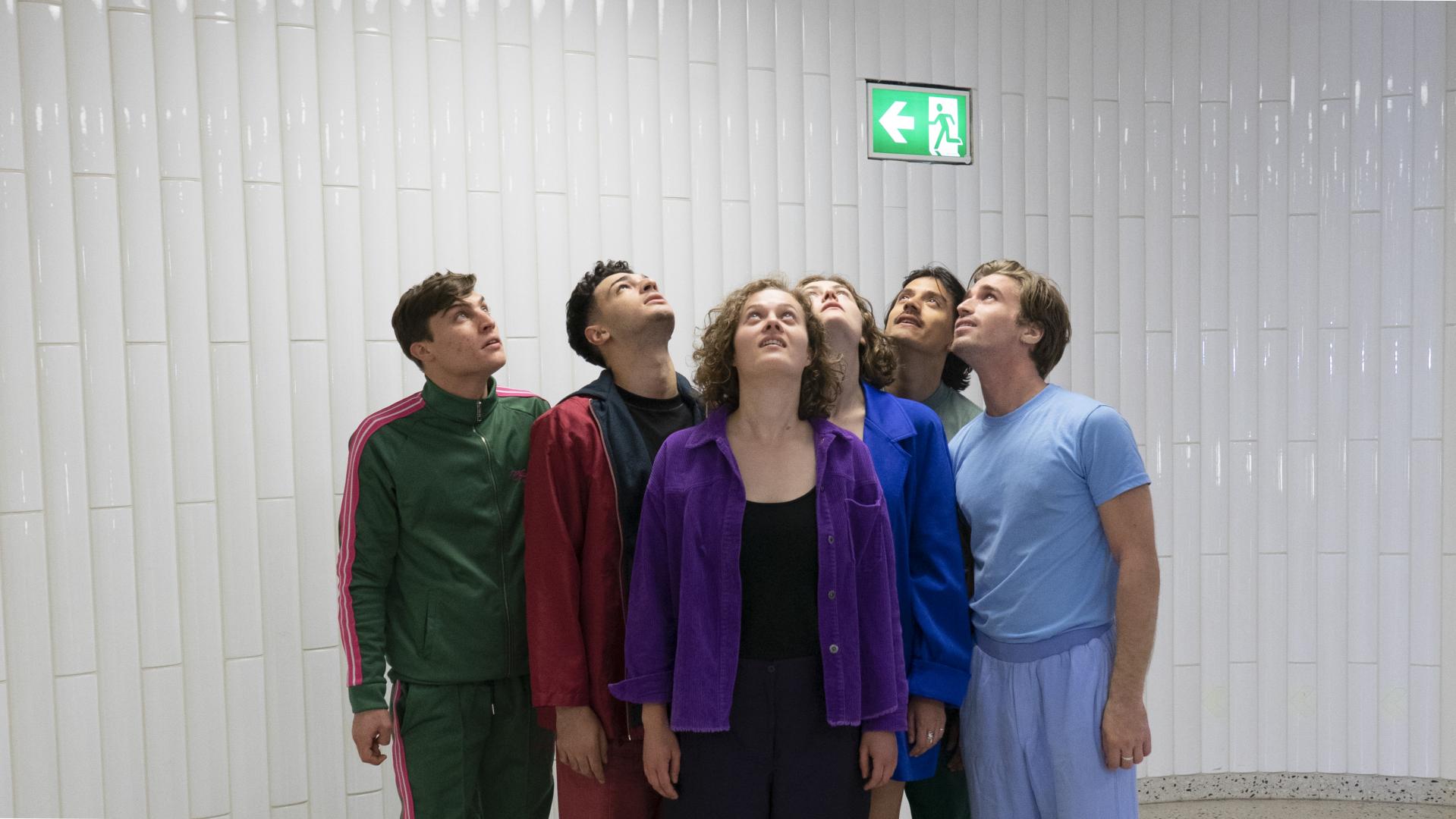 Sechs Schauspielabsolvierende nebeneinander, den Blick nach oben zu einem Notausgangsschild gerichtet.