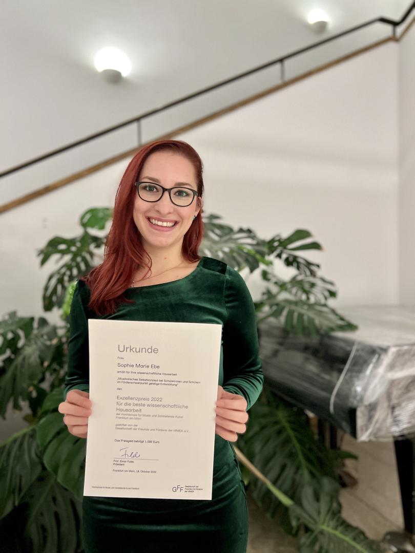 Sophie Marie Ebe im Foyer der HfMDK mit ihrer Urkunde für den Exzellenzpreis der besten wissenschaftlichen Hausarbeit in der Hand