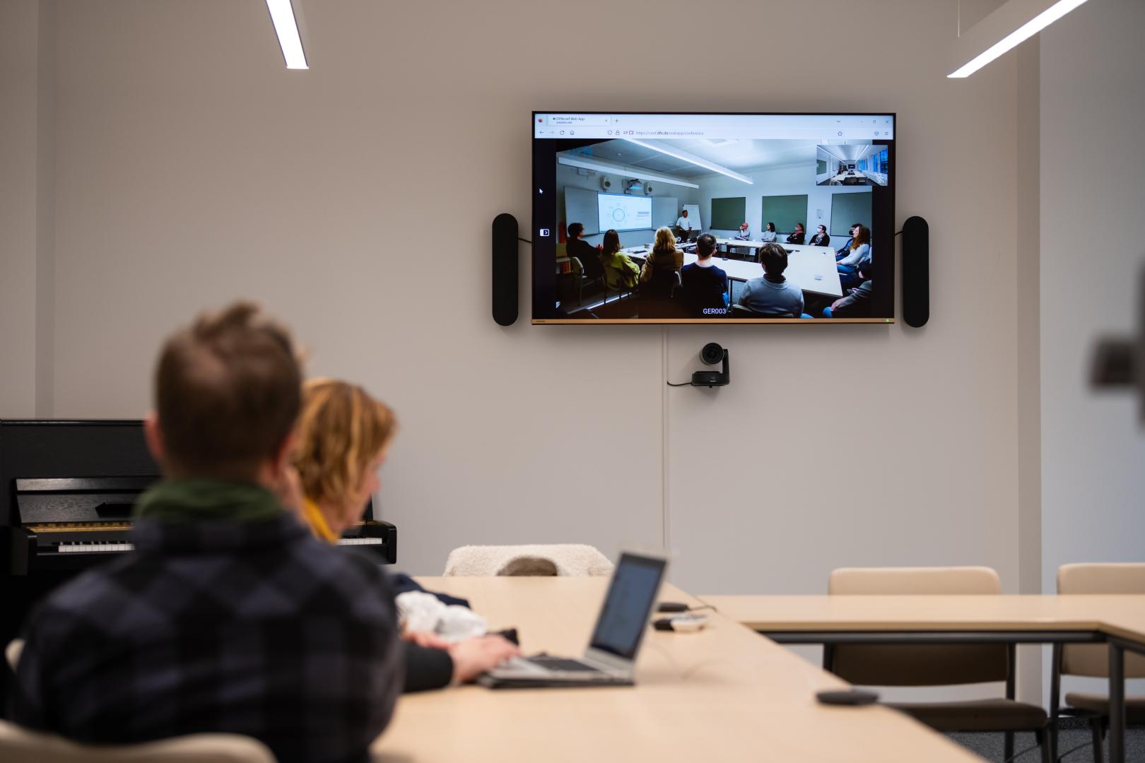 Seminarraum, Fokus des Bilds liegt auf einem Monitor an der Wand, auf den ein Seminar aus einem anderen Raum übertragen wird.