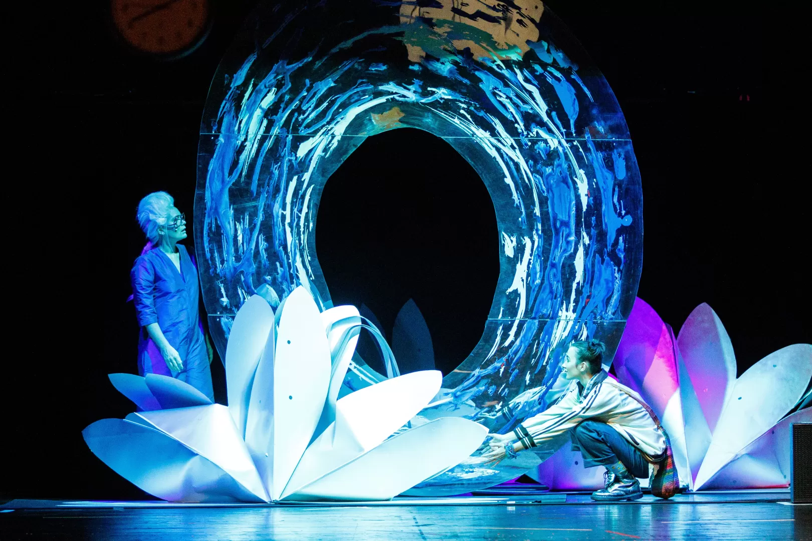 Zwei Schauspielerinnen auf blau beleuchteter Bühne zwischen großen Papierblüten.