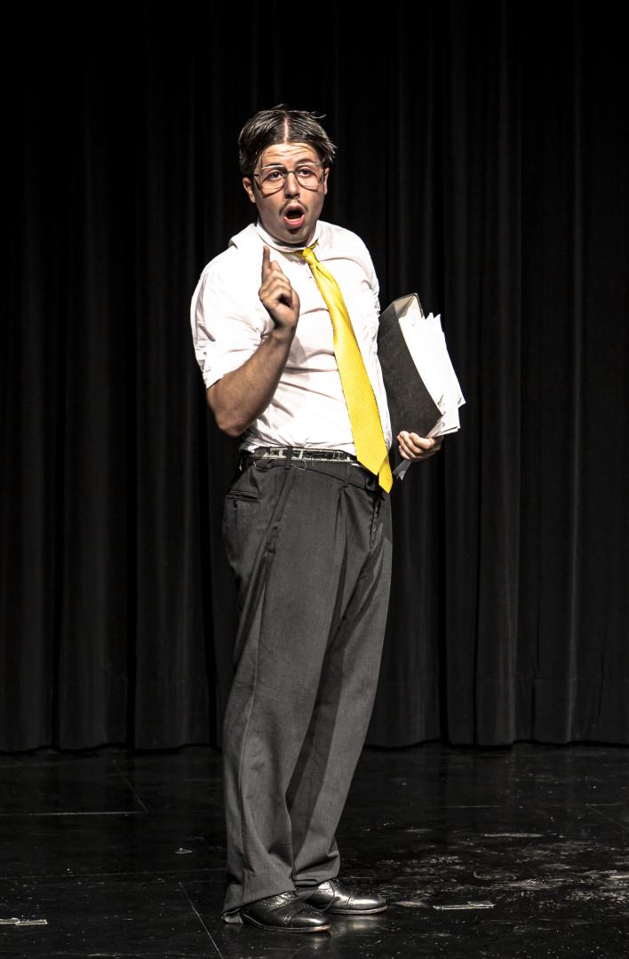 Schauspieler mit Mittelscheitel, Brille, Ordner, gelber Krawatte und erhobenem Zeigefinger.