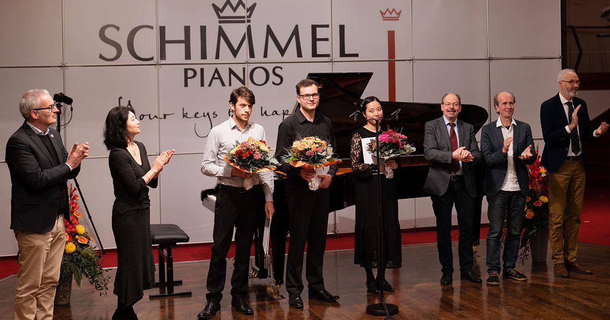 Mehrere Personen stehen auf einer Bühne und klatschen. Drei Personen in der Mitte mit Blumensträußen.