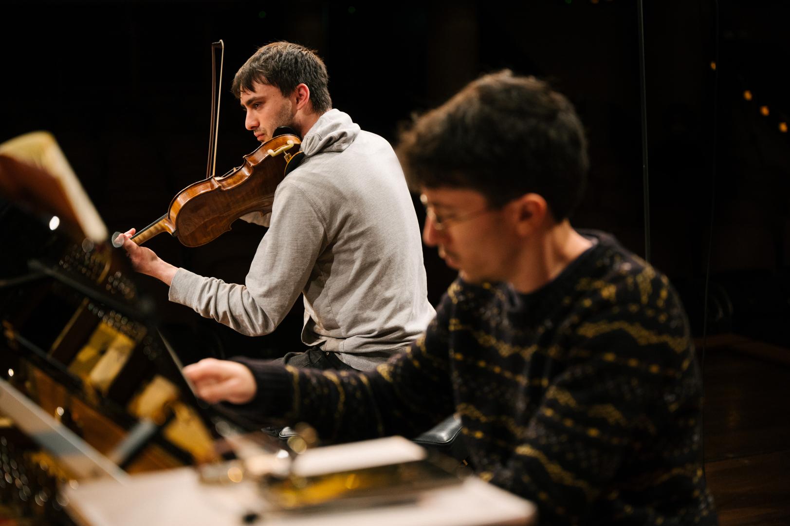 Ein Geiger und ein Pianist während einer gemeinsamen Kammermusikprobe, der Bildfokus liegt auf dem Geiger im Hintergrund des Bildes.