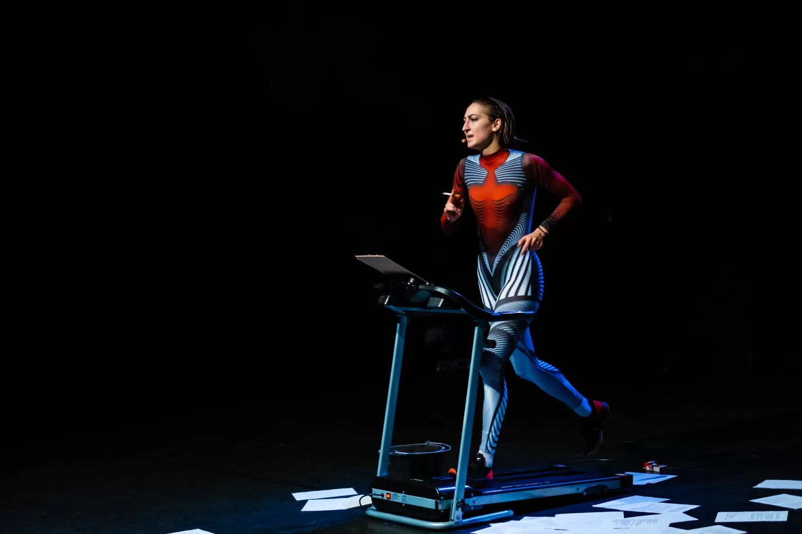 Schauspielerin rennend auf einem Laufband mit einer Zigarette in der Hand und zwischen losen Zetteln auf dem Boden.