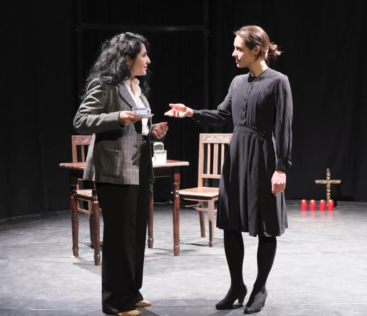 Zwei Frauen auf der Bühne, sie unterhalten sich einander gegenüberstehend. Eine hält eine Teetasse in der Hand.