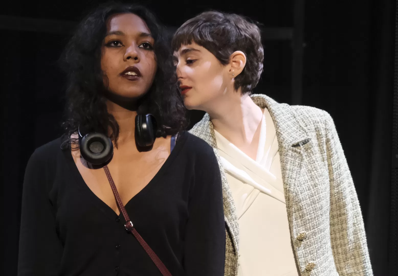 Zwei Frauen in einer Bühnenszene: Eine im hellen Kostüm, die einer anderen Frau in schwarz gekleidet etwas ins Ohr flüstert.