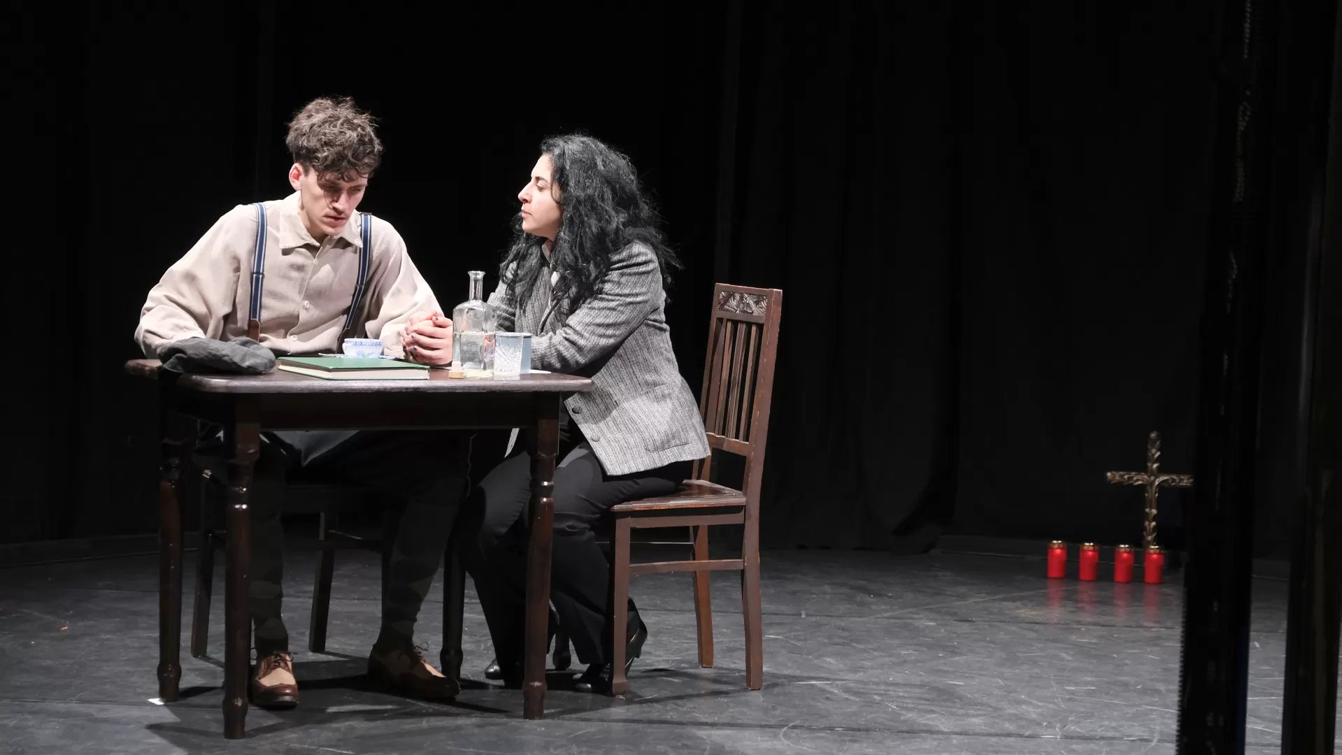 Zwei Personen sitzen auf der Bühne an einem Holztisch, sie halten einander an den Händen.