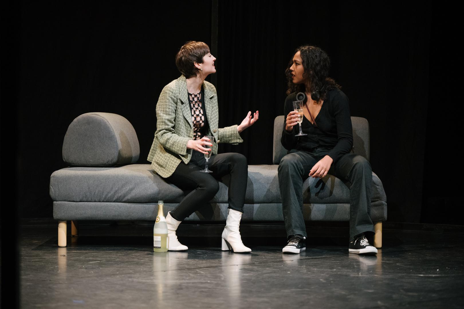 Zwei Schauspielstudentinnen in einer Szene: Sie sitzen auf einem grauen Sofa, einander zugewandt im Gespräch, in den Händen Sektgläser.