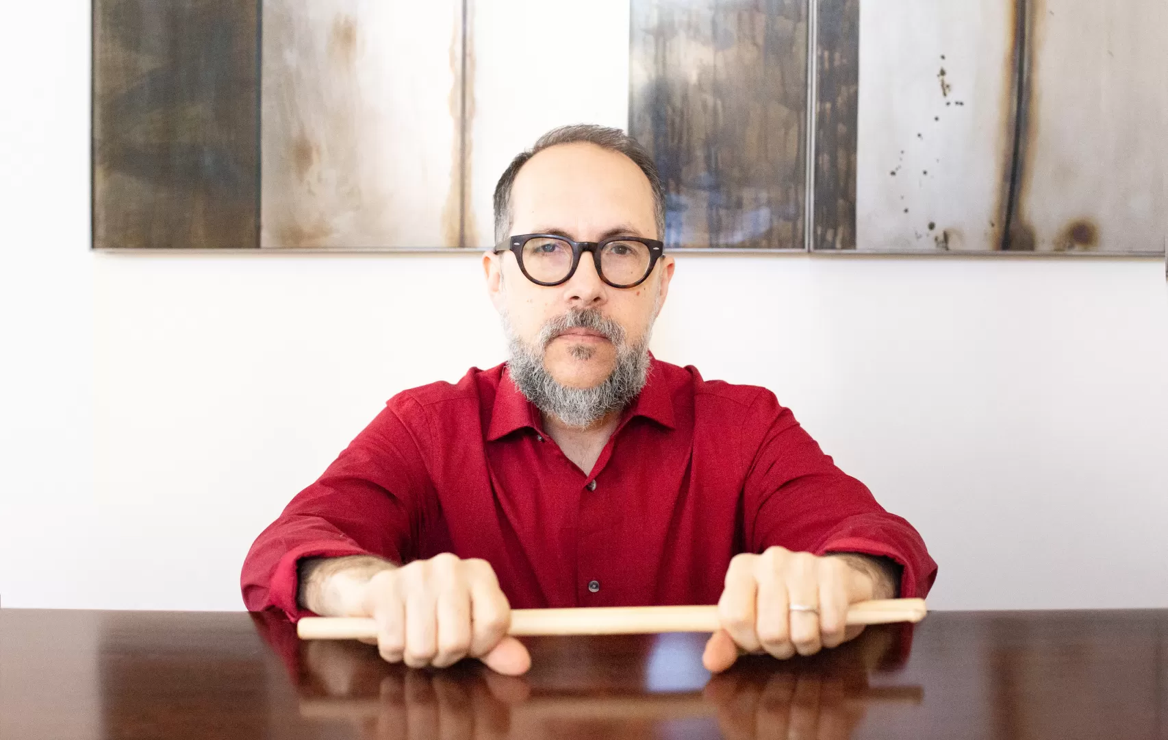 John Hollenbeck sitzt an einem Tisch dem Bildbetrachter gegenüber, in der Hand Schlagzeugsticks.