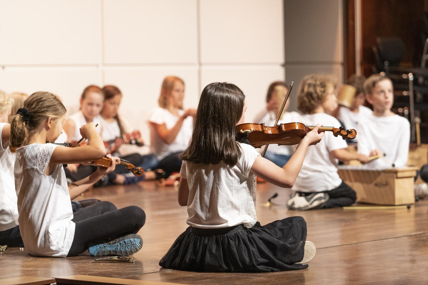 Grundschüler*innen sitzen mit ihren Instrumenten auf der Bühne in der Alten Oper. Im Vordergrund ein Mädchen mit einer Geige in der Hand.