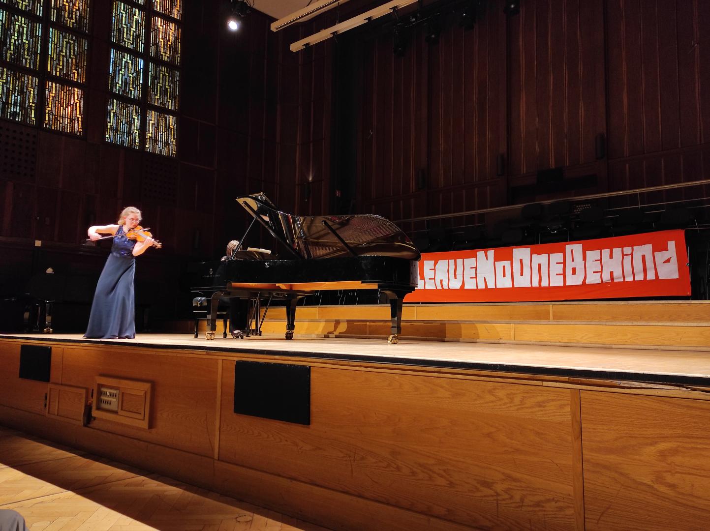 Konzert im Großen Saal der HfMDK. Auf der Bühne ein Flügel und eine Geigerin, hinten am Bühnenrand hängt ein Transparent mit der Aufschrift Leave No One Behind.