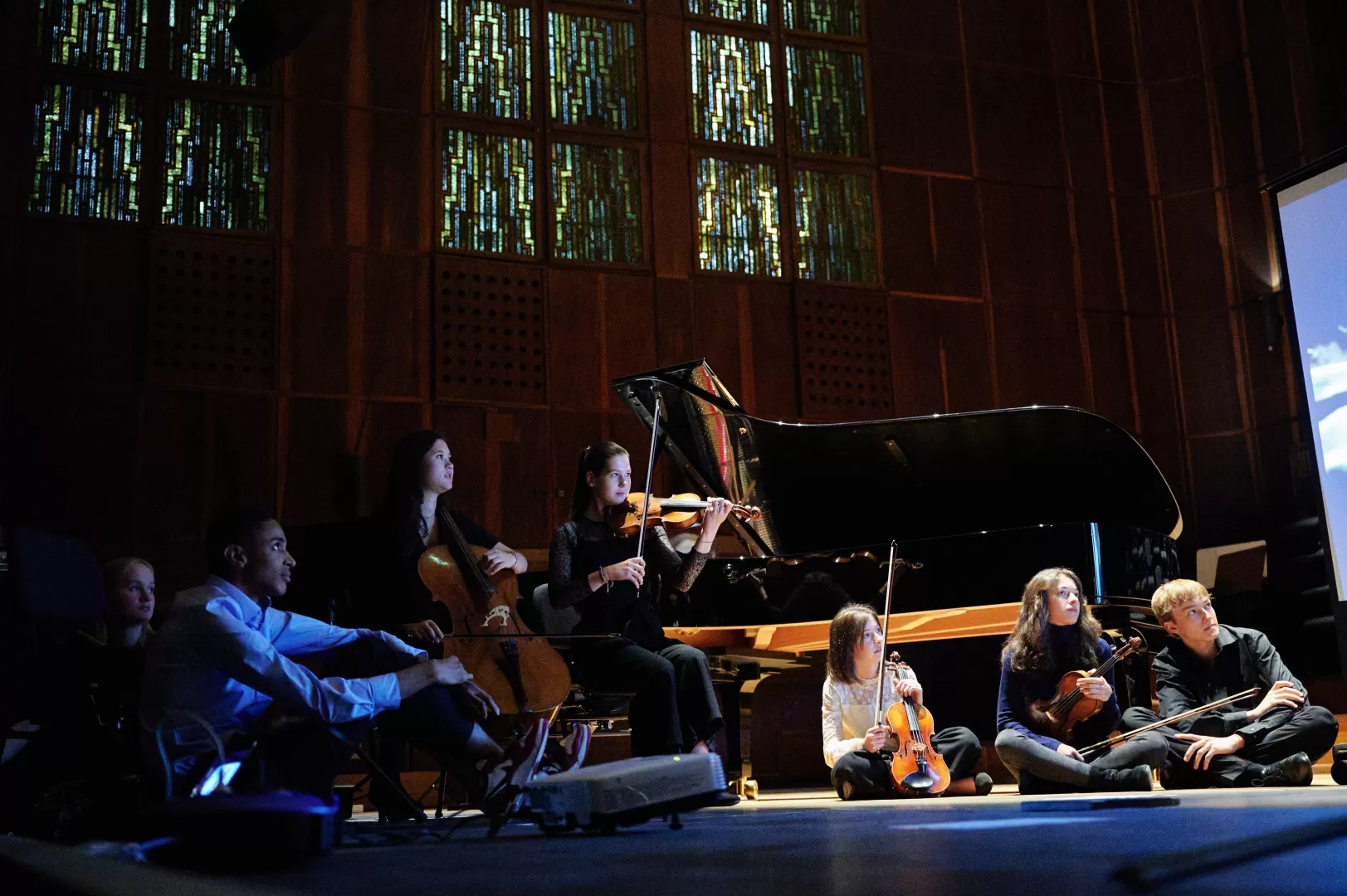 Mehrere Personen sitzen im Halbdunkeln und blicken in eine Richtung. Eine Person spielt Geige, eine weitere spielt Violoncello..