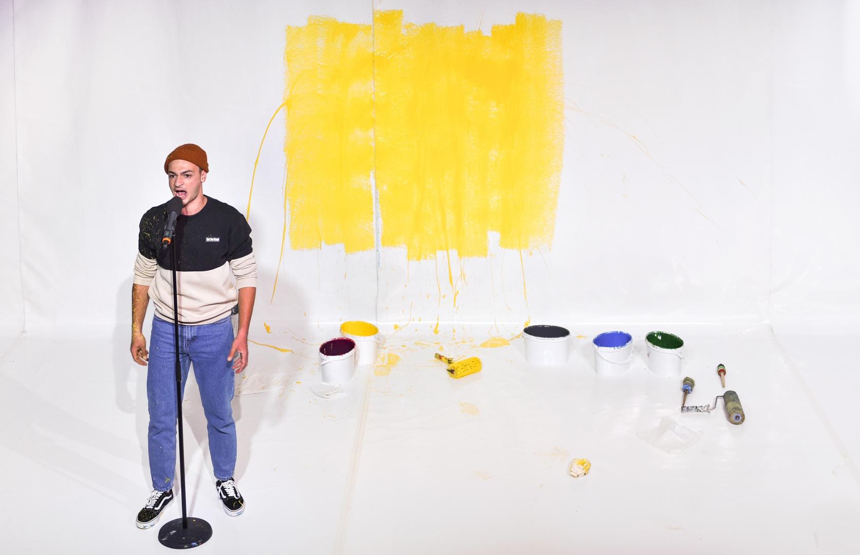 Schauspieler steht vor an einem Stehmikrofon vor einer weißen Wand, davor liegen Farbeimer und -rollen.