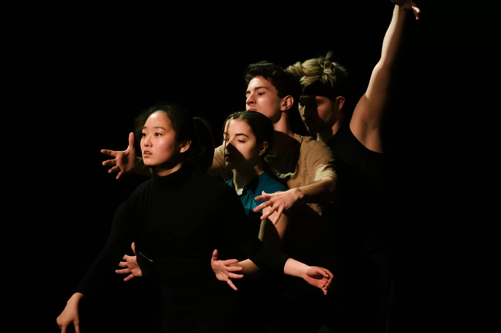 Vier Tänzer*innen stehen hintereinander, sie sind dunkel gekleidet und strecken ihre Arme in unterschiedliche Richtungen aus.