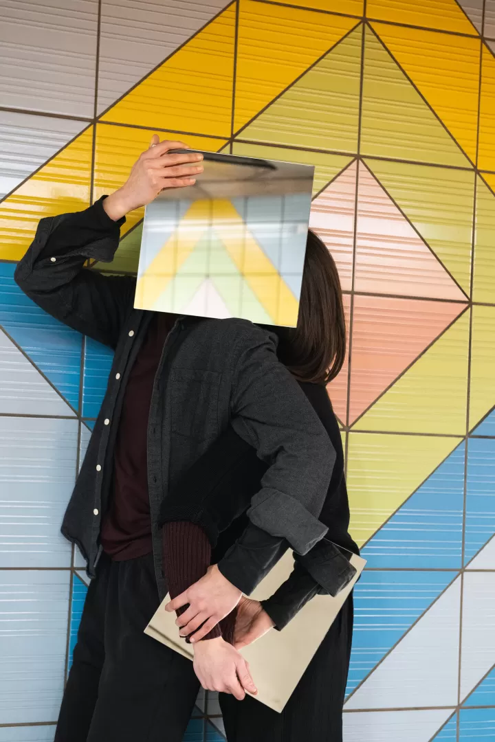 Zwei Personen in schwarz gekleidet, sie stehen eng beieinander, die Gesichter mit quadratischen Spiegeln verdeckt, die die umliegenden bunten Kacheln der U-Bahnstation Grüneburgweg reflektieren.