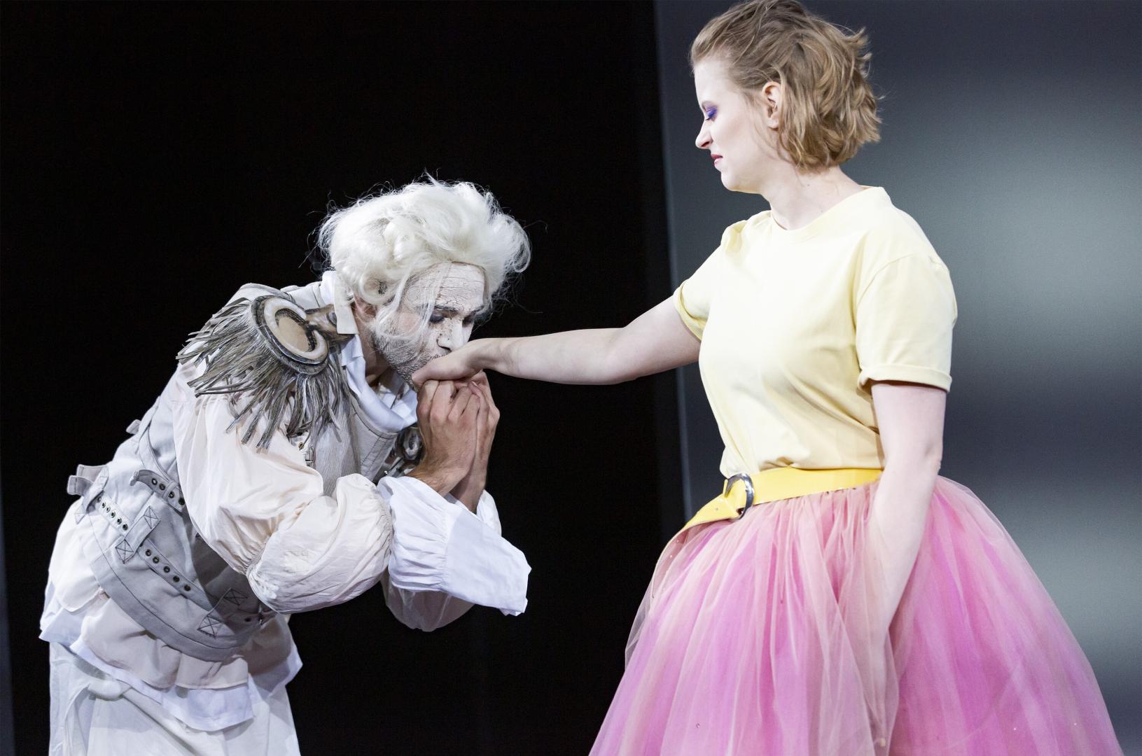 Schauspieler mit gepuderter Perücke gibt einer Schauspielerin, die angewidert schaut, einen Handkuss.