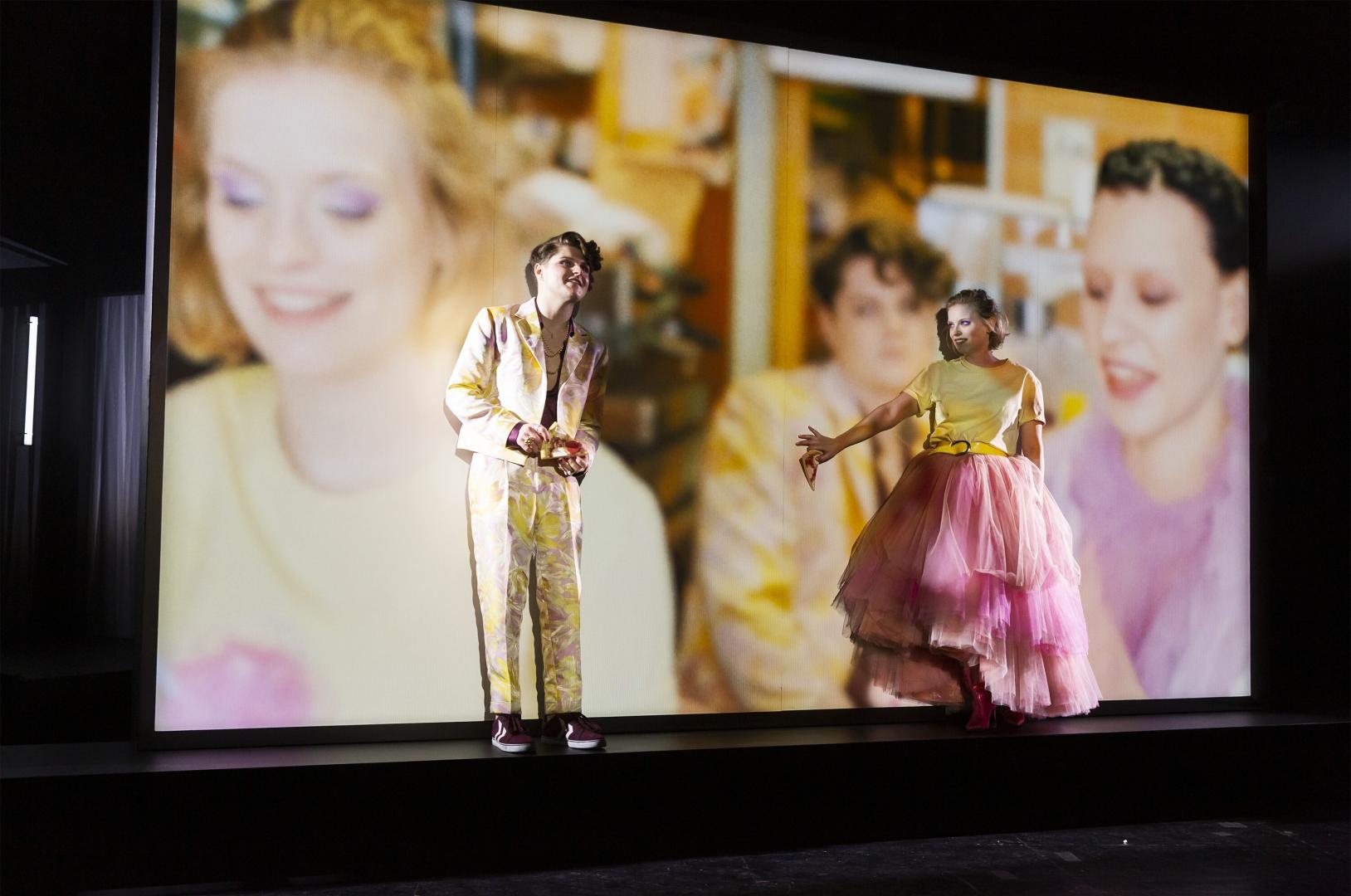 Zwei Schauspielende vor einer Videoleinwand schauen verträumt.