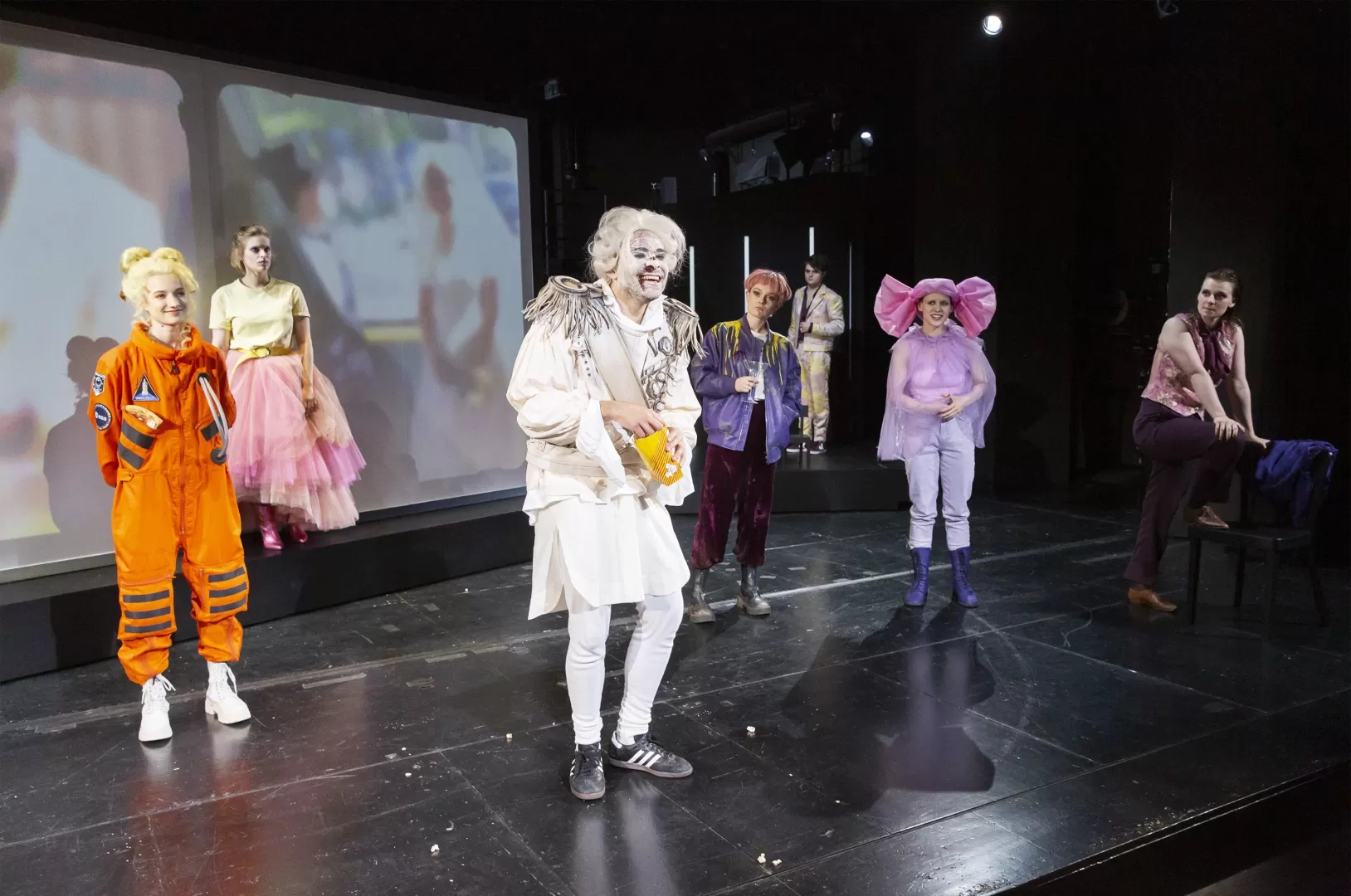 Sieben bunt kostümierte Schauspielende, in der Mitte ein Mann mit gepuderter Perücke, zu dem alle hinsehen.