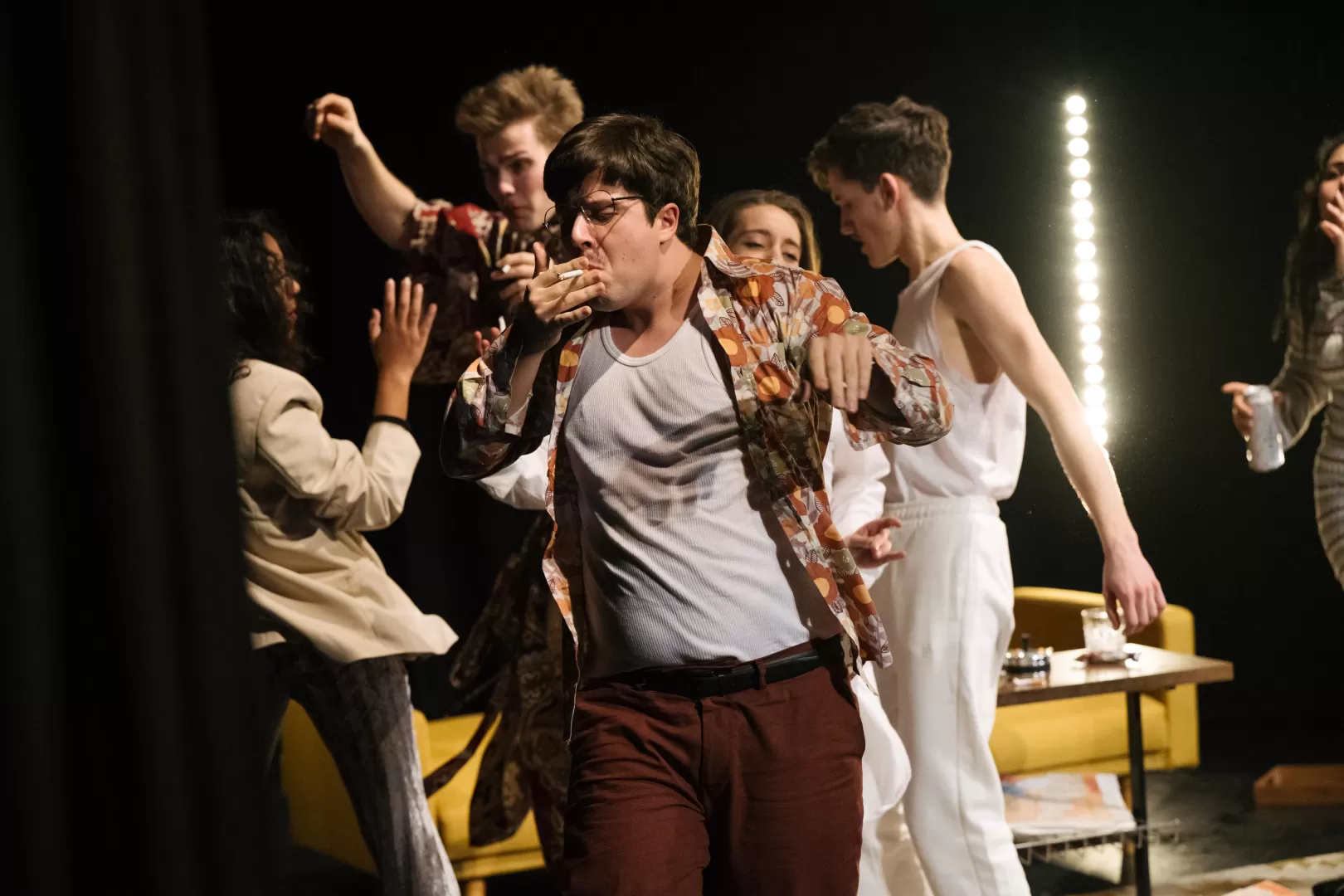 Mehre Schauspielende tanzen vor einem gelben Sofa, ein Schauspieler im Vordergrund mit Zigarette in der Hand.