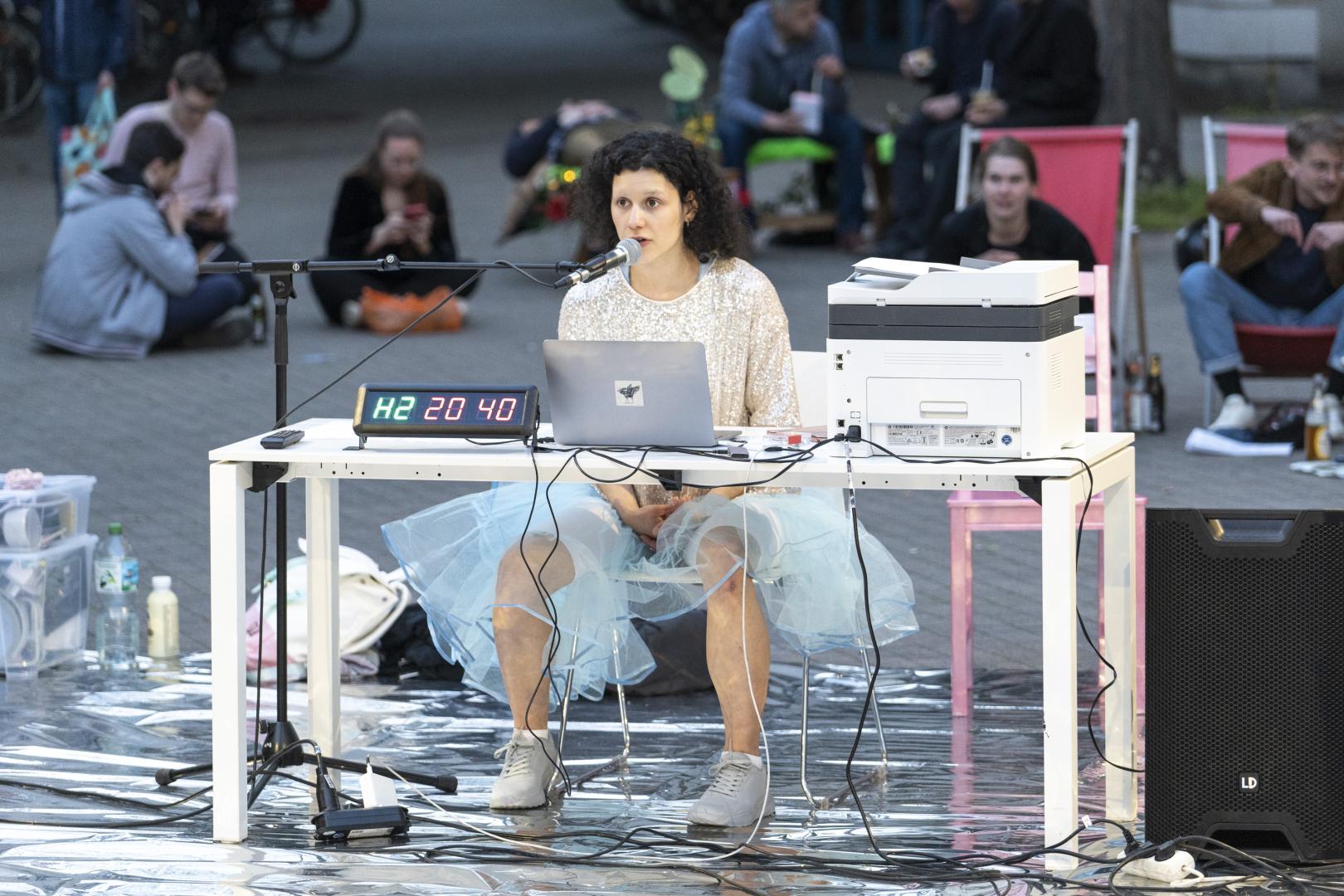 Szene aus einer Performance im Innenhof der HfMDK: Eine Darstellerin sitzt vor einem Laptop an einem Tisch und spricht ins Mikrofon