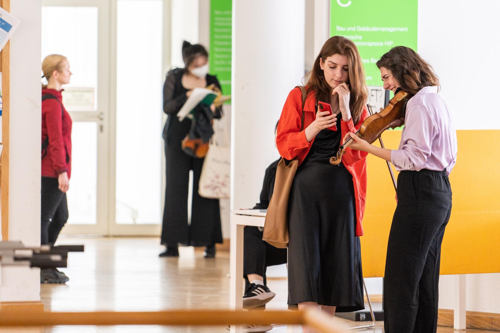 Zwei Studentinnen, von denen eine eine Geige in der Hand hält, beugen sich über ein Smartphone.