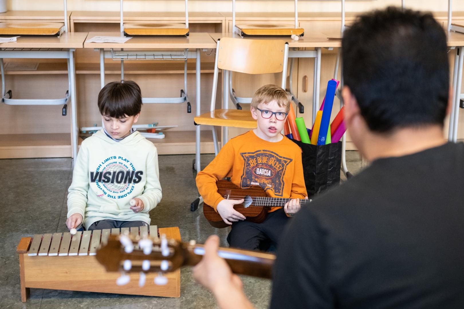 Zwei Kinder mit Musikinstrumenten, vor ihnen ein Student mit einer Gitarre.