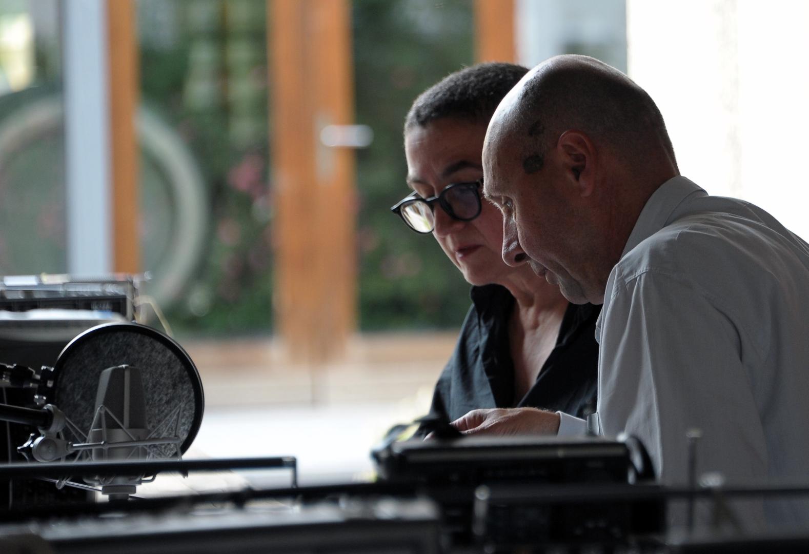 Annette Schmucki und Reto Friedmann sitzen an einem Tisch mit Mikrofonen und anderen technischen Geräten.