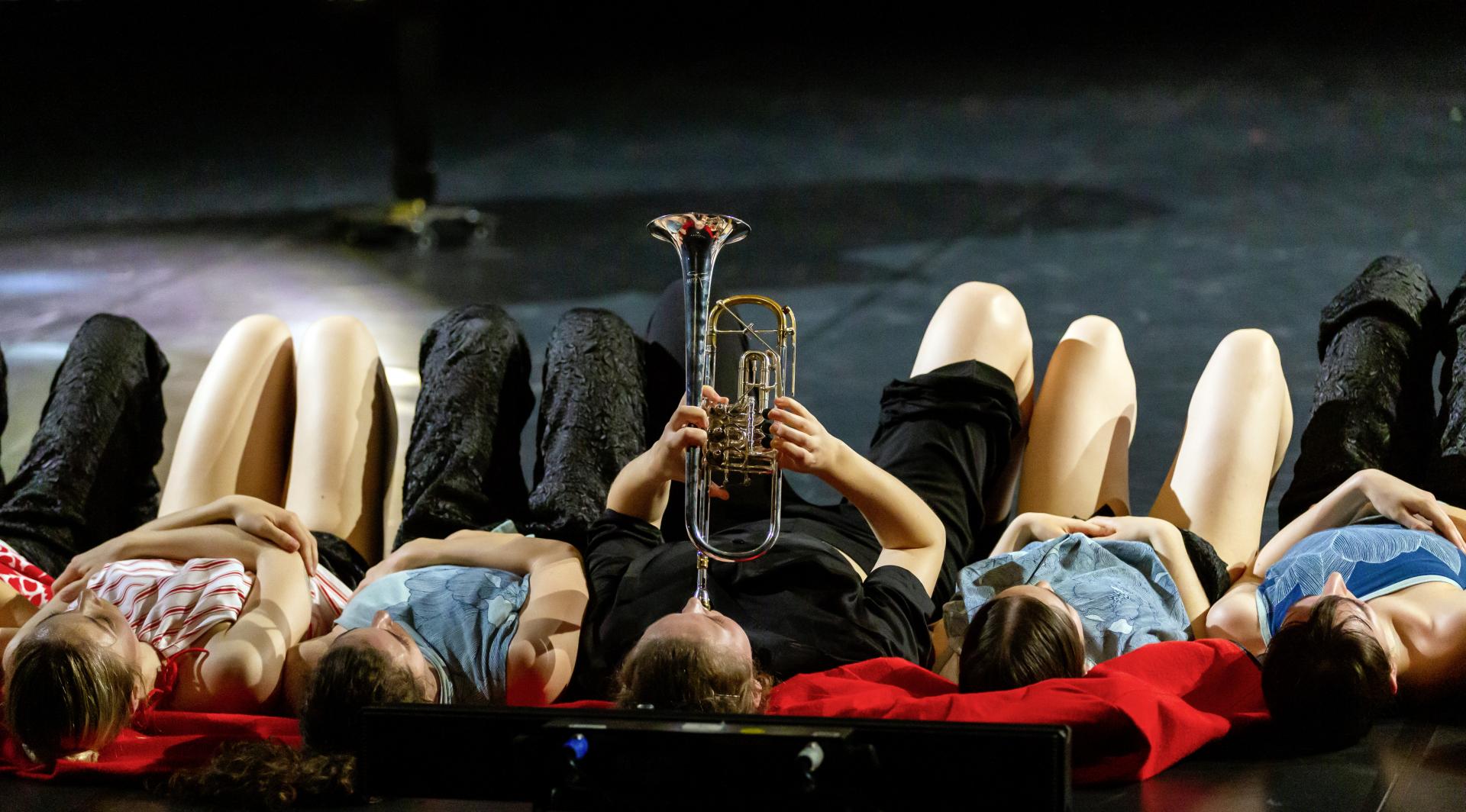 Fünf Personen liegen auf der Bühne auf dem Boden. Die Person in der Mitte spielt Trompete.