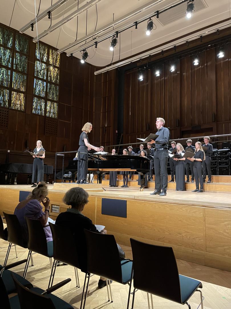 Studentin dirigiert Chor, zwei Solist*innen singen vorne an der Bühne